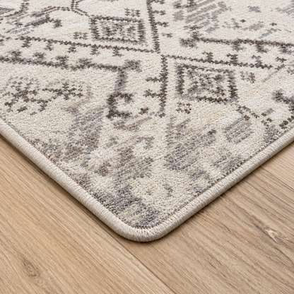 Afterimage area rug: close-up of a patterned area rug corner on a light wood floor. The rug has a cream and gray traditional-style design with a thick beige border.