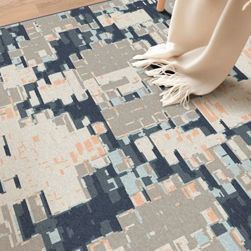 A modern abstract area rug with a geometric block pattern in navy, gray, beige, and light blue, styled in a room setting with a cream throw blanket draped over a wooden chair.