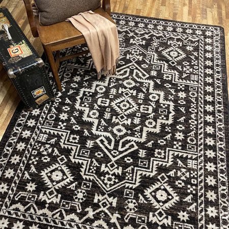 A rectangular area rug with a brown and off-white geometric tribal pattern, styled on a wooden floor next to a wooden chair with a beige throw blanket and a vintage trunk.