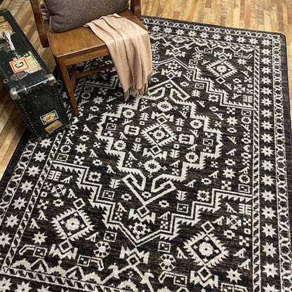 A rectangular area rug with a brown and off-white geometric tribal pattern, styled on a wooden floor next to a wooden chair with a beige throw blanket and a vintage trunk.