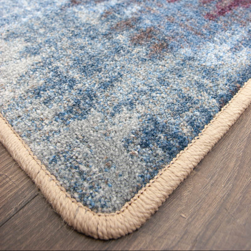 Color Rush area rug: close-up of a ttextured area rug corner showing a blue, gray, and red abstract pattern with a thick beige braided border on a dark wood floor.