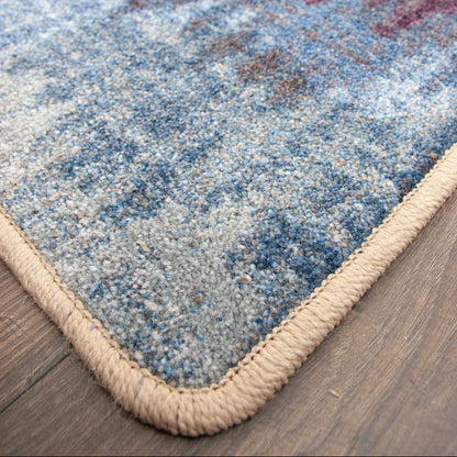 Color Rush area rug: close-up of a ttextured area rug corner showing a blue, gray, and red abstract pattern with a thick beige braided border on a dark wood floor.