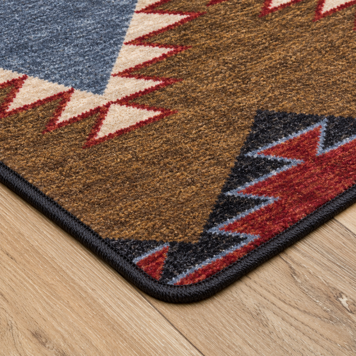 Crossplay area rug: close-up of a patterned area rug corner on a light wood floor. The rug has a brown field with geometric Southwestern-style motifs in blue, cream, red, black, and gray, finished with a…