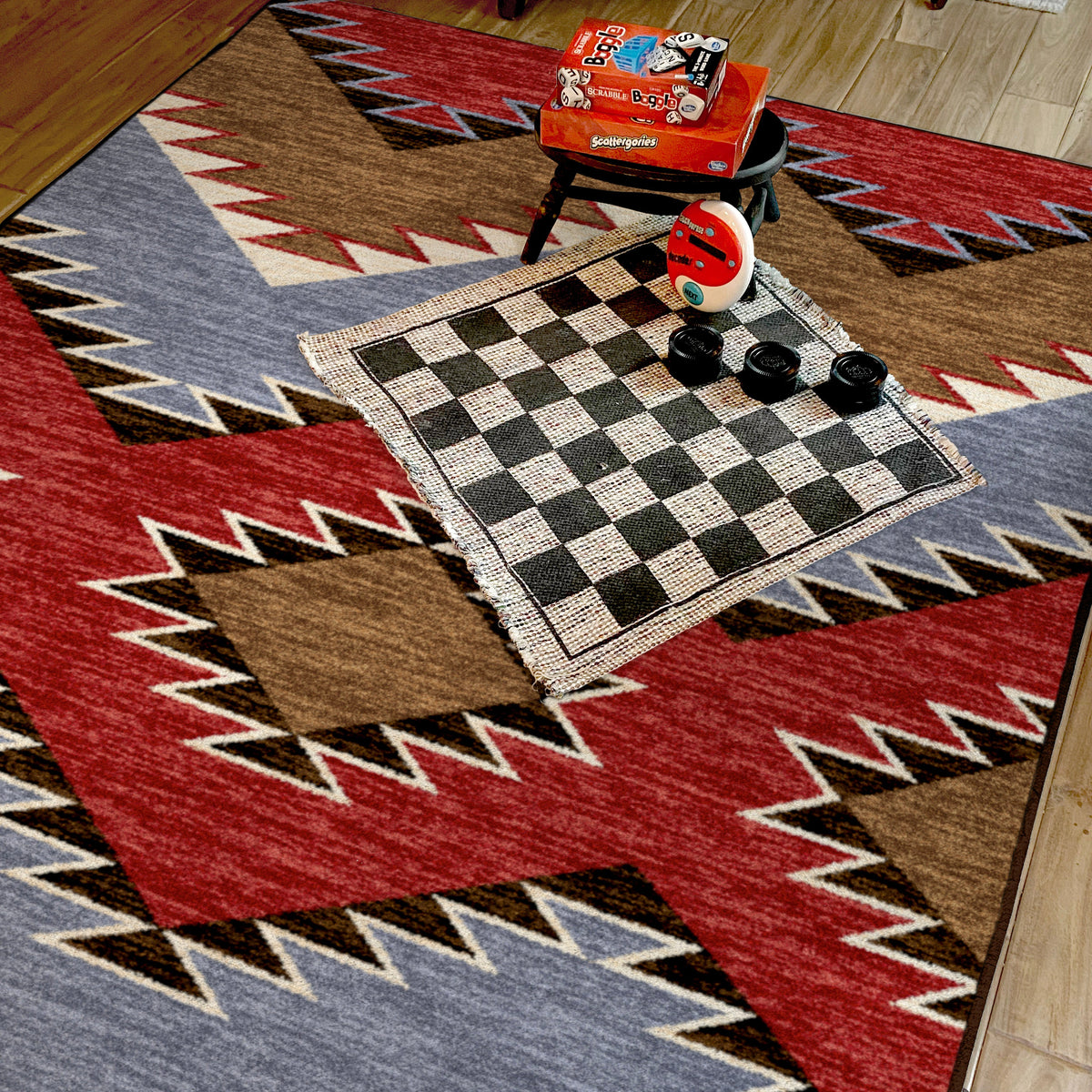 Crossplay area rug: a Southwestern-style area rug with a bold geometric pattern in red, brown, slate blue, cream, and black, styled in a room setting with a small black stool, a red box, and a checkered…