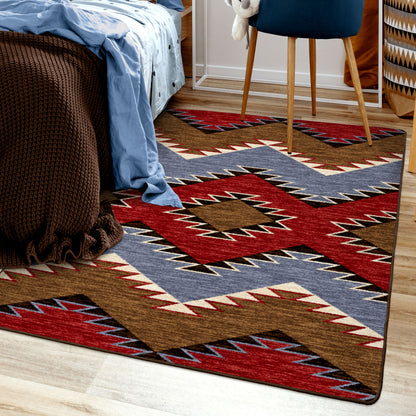 Crossplay area rug: a rectangular area rug with a Southwestern geometric pattern in red, brown, blue, beige, and black, shown on a light wood floor in a bedroom setting next to a bed and chair.