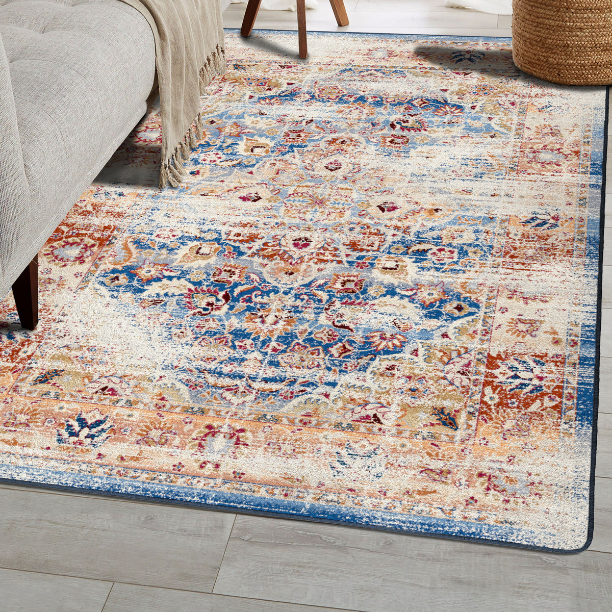 A rectangular area rug with a distressed traditional pattern in blue, beige, rust, and cream, shown on a light wood floor next to a gray upholstered chair and a woven basket.