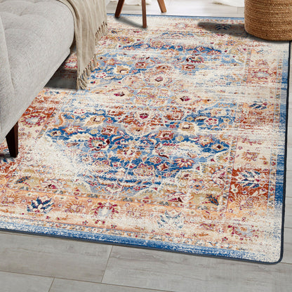 A rectangular area rug with a distressed traditional pattern in blue, beige, rust, and cream, shown on a light wood floor next to a gray upholstered chair and a woven basket.