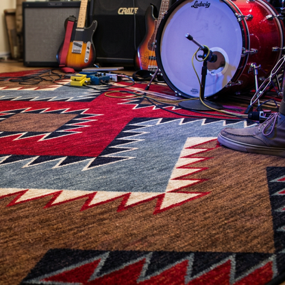 A Southwestern-style patterned area rug in red, blue, brown, and beige is shown on a wooden floor in a music room setting, with parts of a person's legs and shoes visible at the edges, along with guitars…