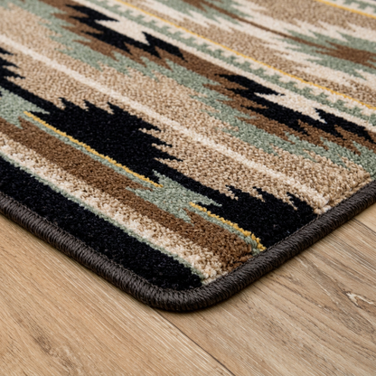 Field Notes area rug: a close-up of a patterned area rug corner on a light wood floor. The rug has a Southwestern-style geometric design in beige, brown, black, sage green, and muted yellow, finished with a…
