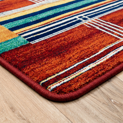 Framework area rug: a close-up of a colorful area rug corner on a light wood floor, showing a geometric pattern in rust orange, red, teal, yellow, and navy with a thick red border.