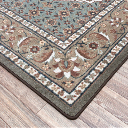 A close-up corner view of a traditional area rug with an ornate floral and scroll pattern, shown on a light brown wood floor. The rug features a muted blue-gray field, brown and beige borders, and dark brown edges.