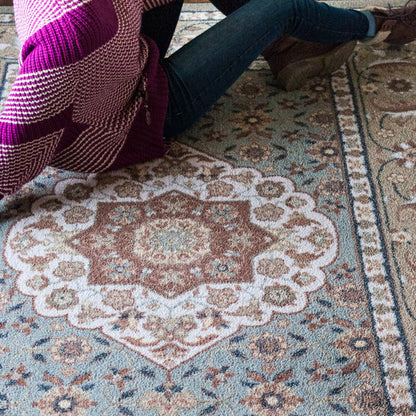A close-up of a patterned area rug with a central medallion design in muted blue, beige, brown, and pink tones. A person wearing blue jeans and brown boots is sitting on the rug, with a pink and white knit blanket draped over their lap.