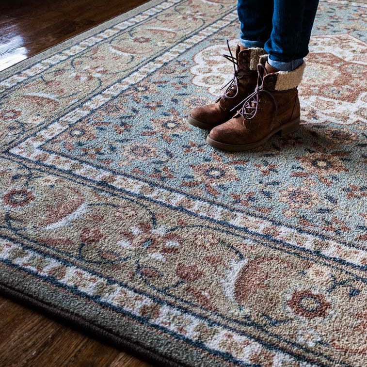 A close-up of a traditional patterned area rug on a dark wood floor, with a person's legs and brown lace-up boots visible standing on the rug.