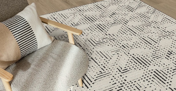 A close-up of a cream and black geometric patterned area rug styled in a modern living room setting next to a light wood armchair with a ttextured throw blanket.