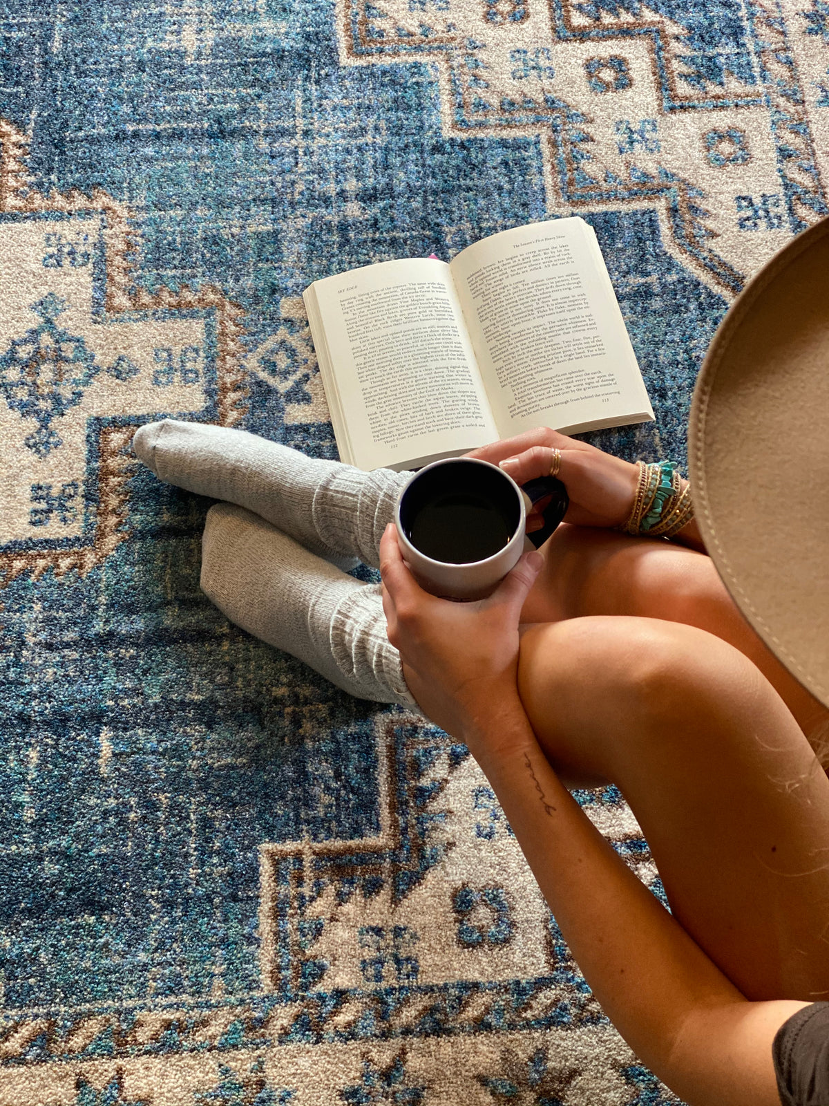 A person sitting on a blue and beige patterned area rug, holding a black coffee mug and an open book. The person is wearing light gray socks, bracelets, and a tan wide-brim hat.