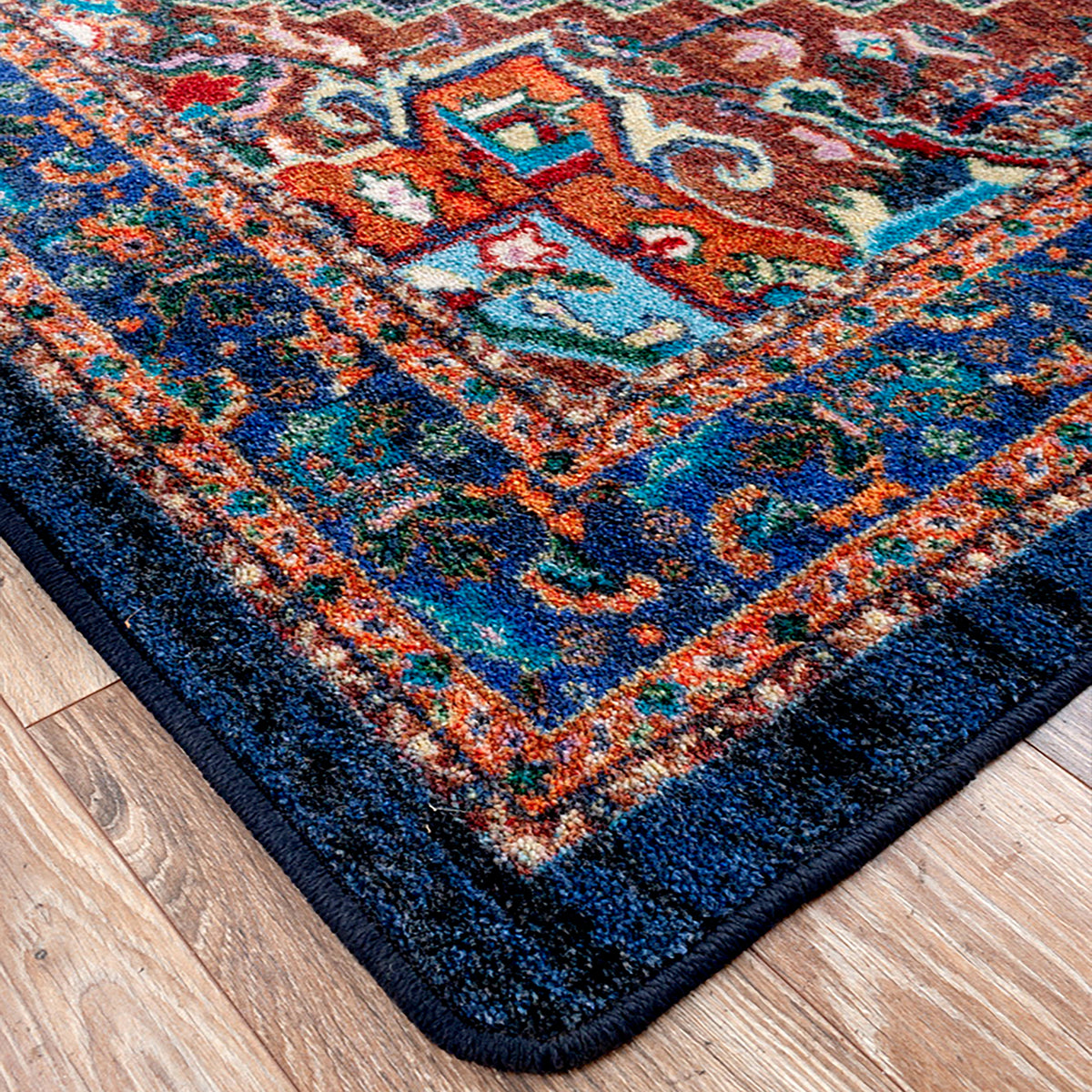 Good Company area rug: a close-up corner view of a traditional patterned area rug with a dark navy blue border and a multicolor ornamental field, shown on a light wood floor.