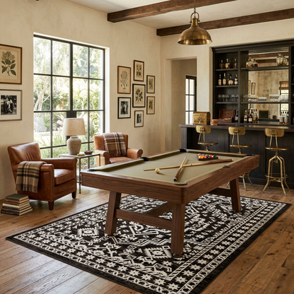 A wooden pool table with a green felt surface is staged in a rustic living room with wood floors, a black-and-white patterned rug, leather armchairs, framed wall art, and a dark bar area in the background.