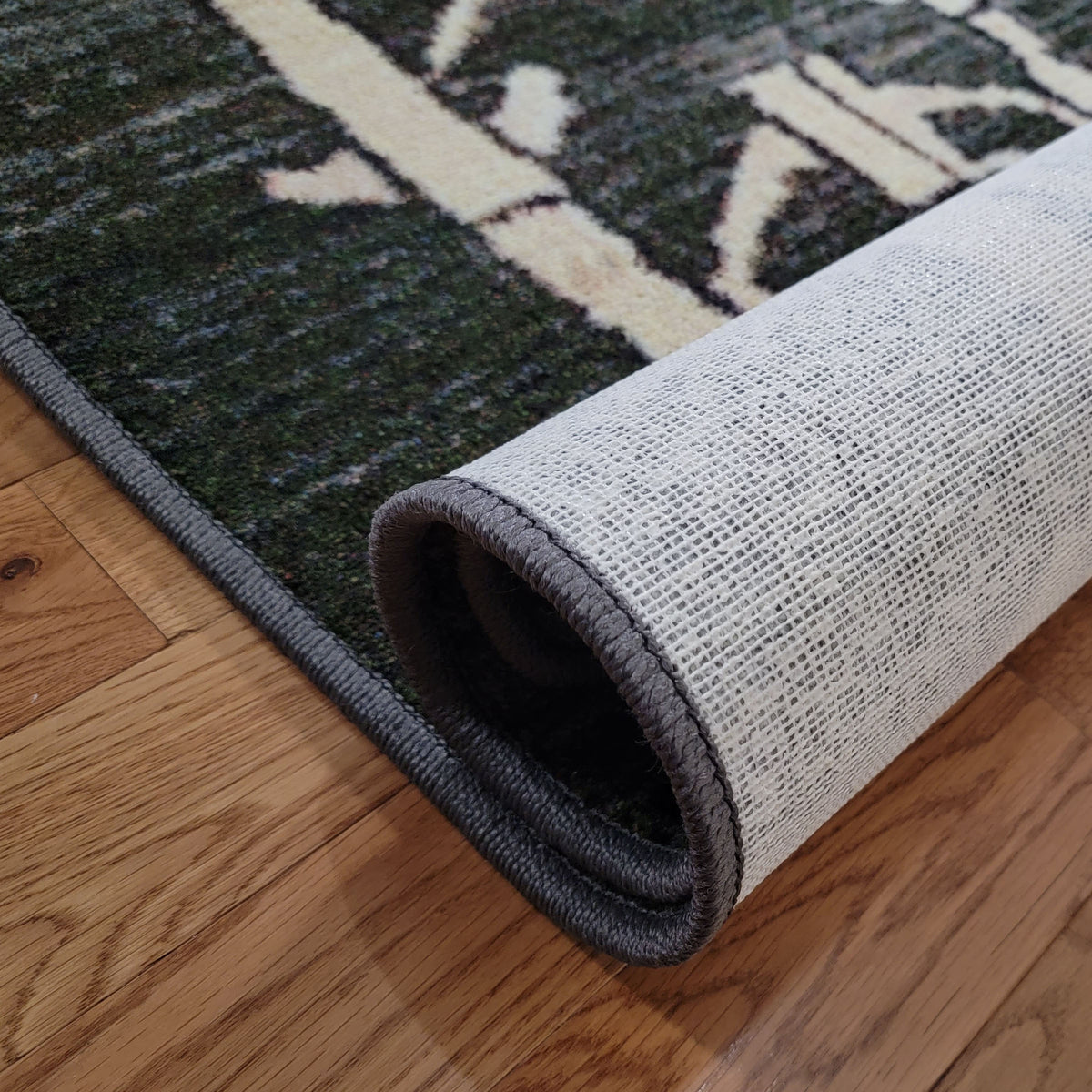 A close-up of a rolled area rug showing its textured cream and dark green patterned top and light gray underside, placed on a hardwood floor.