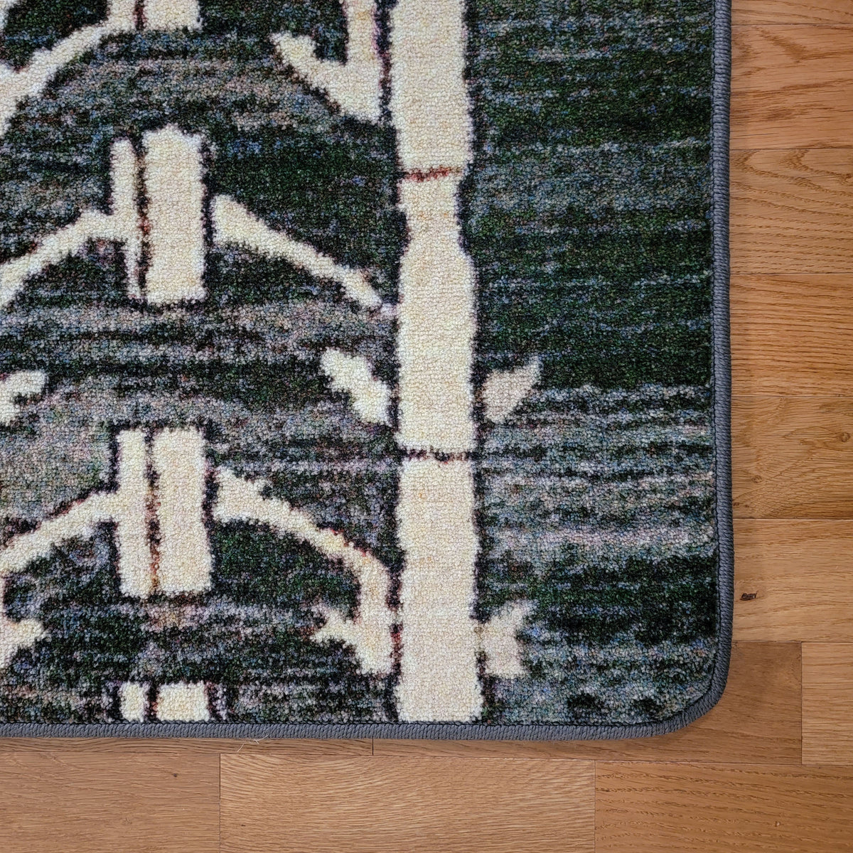 A close-up of a patterned area rug corner on a light wood floor. The rug has a dark green and gray abstract background with large off-white vertical tree-like shapes and a gray border.