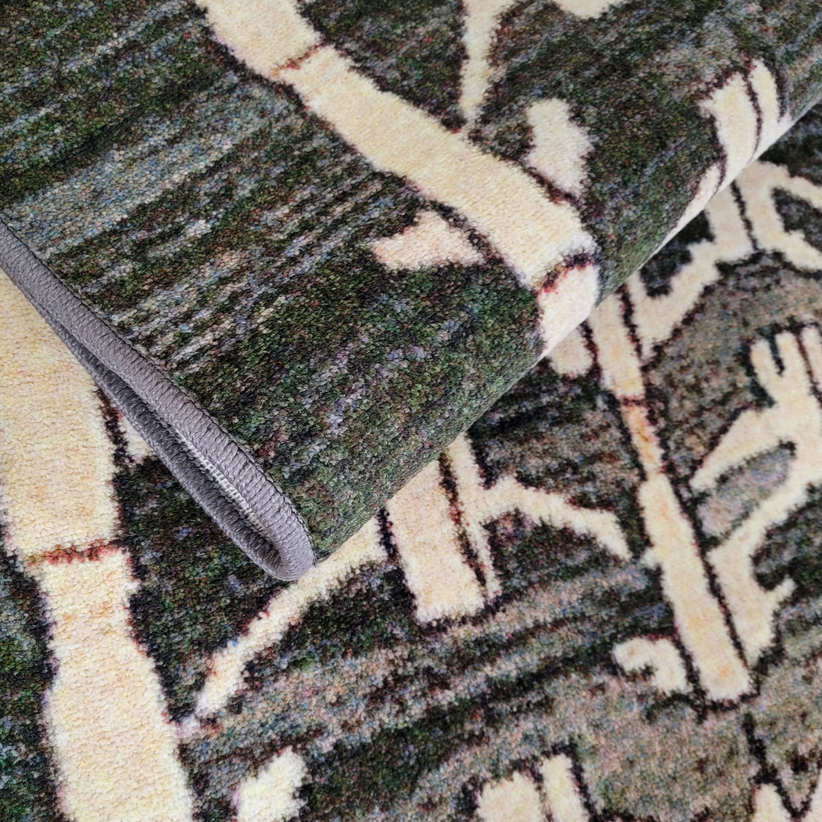 A close-up of a folded area rug showing its textured pile and pattern. The rug features a dark forest green base with cream-colored abstract branch or leaf motifs outlined in black, with a gray binding edge visible on the folded underside.