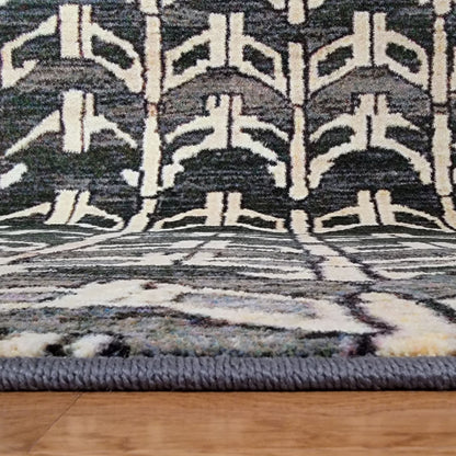 A close-up of a patterned area rug with a thick braided gray border, featuring a geometric design in charcoal, cream, and muted blue-gray, shown on a light wood floor.