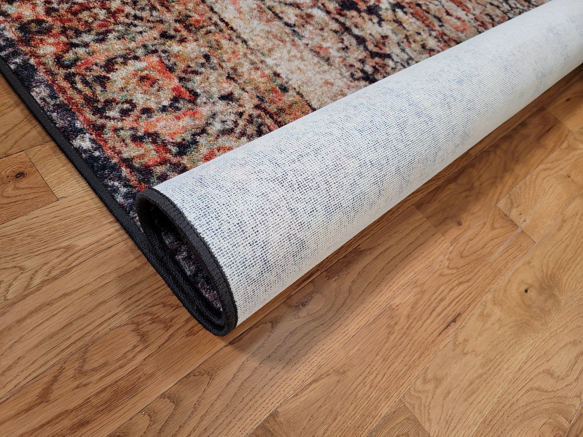 A partially unrolled area rug with a traditional multicolor pattern and a black border, shown on a light brown hardwood floor.