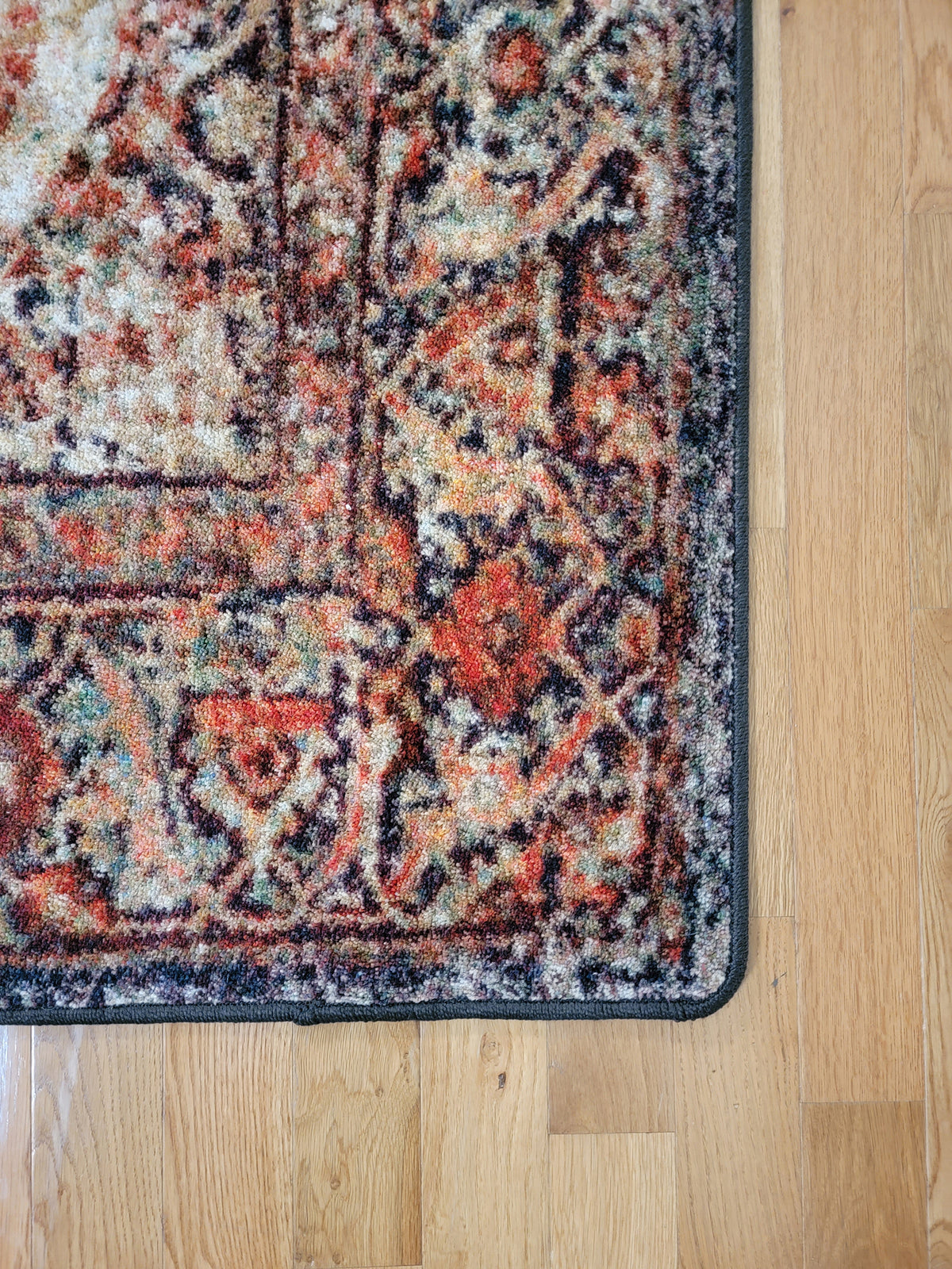 A close-up corner view of a traditional patterned area rug with a distressed design in rust red, beige, and black, shown on a light wood floor.