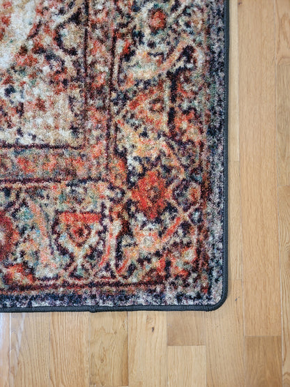 A close-up corner view of a traditional patterned area rug with a distressed design in rust red, beige, and black, shown on a light wood floor.