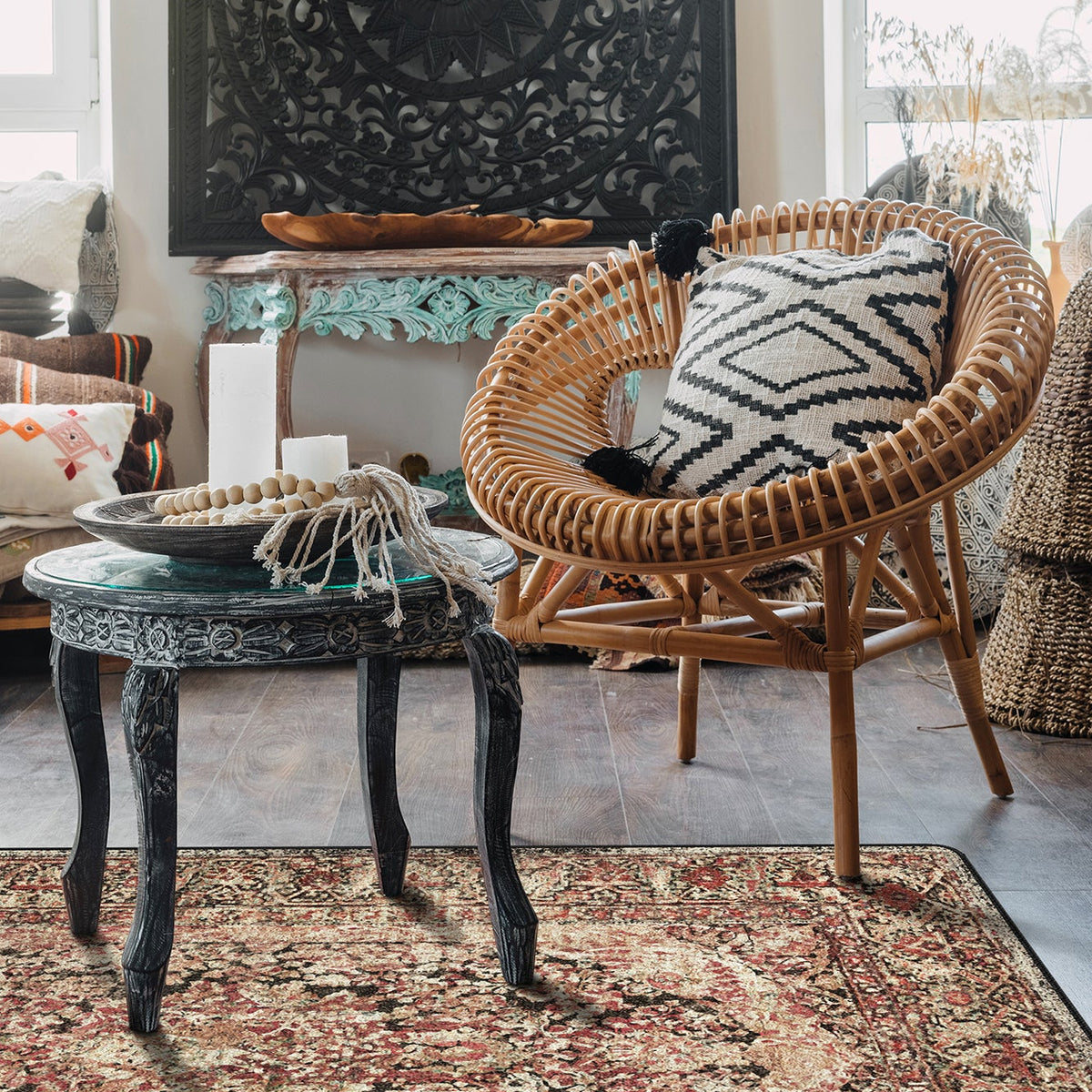 A bohemian-style living room scene featuring a round woven rattan armchair with a black and white geometric patterned throw pillow, set next to a small carved wooden table and a large patterned area rug.