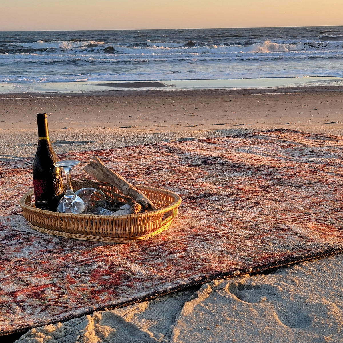 A woven wicker tray holding a dark wine bottle, a wine glass, and driftwood sits on a patterned beach blanket on a sandy beach with the ocean and sky in the background at sunset.