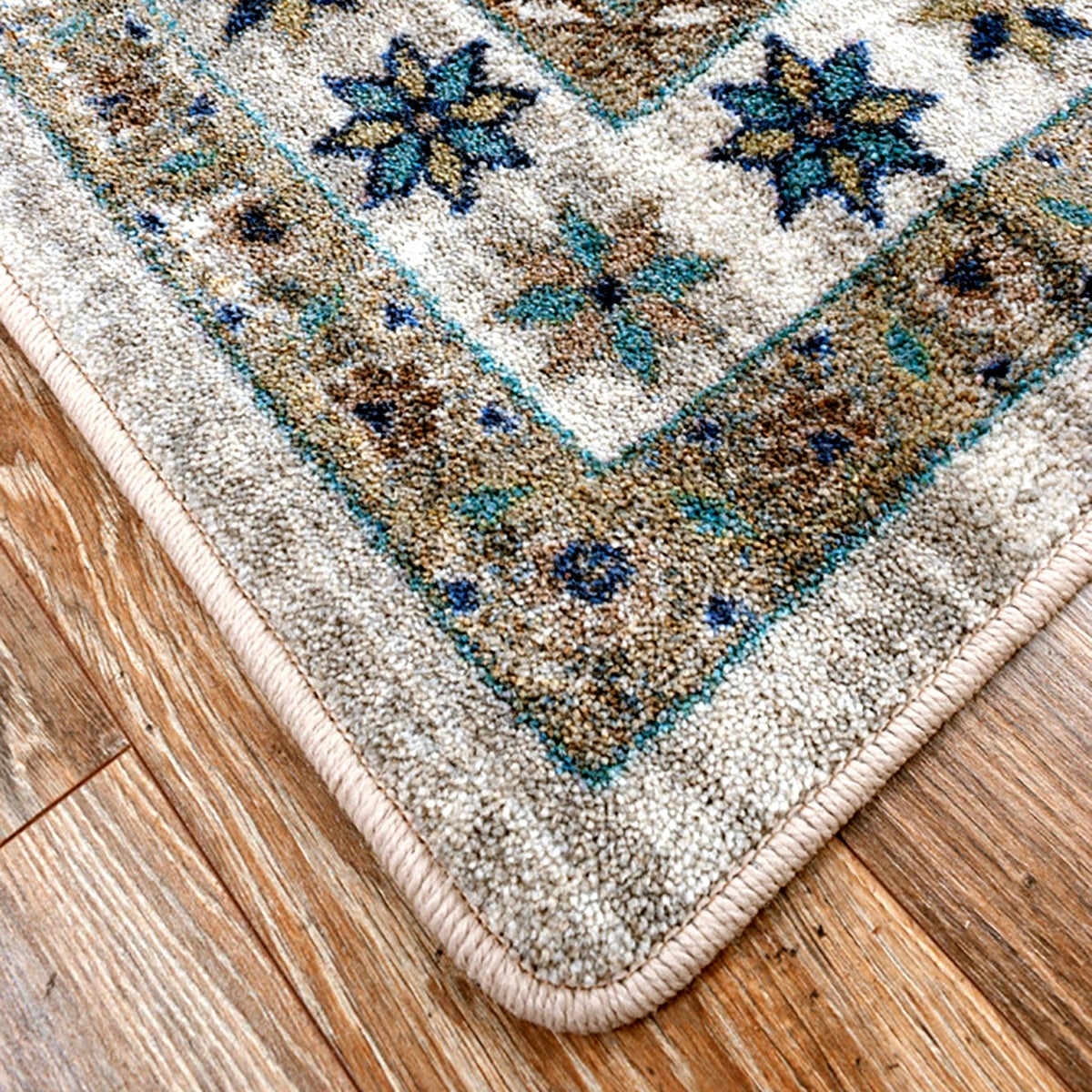 A close-up corner view of a patterned area rug laid on a hardwood floor. The rug has a cream and beige base with blue, teal, and brown ornamental floral and geometric motifs, and a thick cream border.