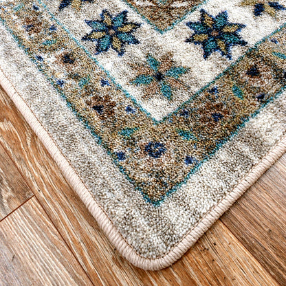 A close-up corner view of a patterned area rug laid on a hardwood floor. The rug has a cream and beige base with blue, teal, and brown ornamental floral and geometric motifs, and a thick cream border.
