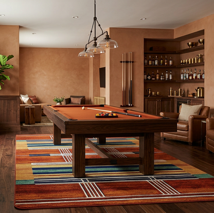 A warm, upscale interior room featuring a wooden ppool table centered on a colorful striped rug, with leather armchairs, built-in shelving, framed wall art, and wood paneling.