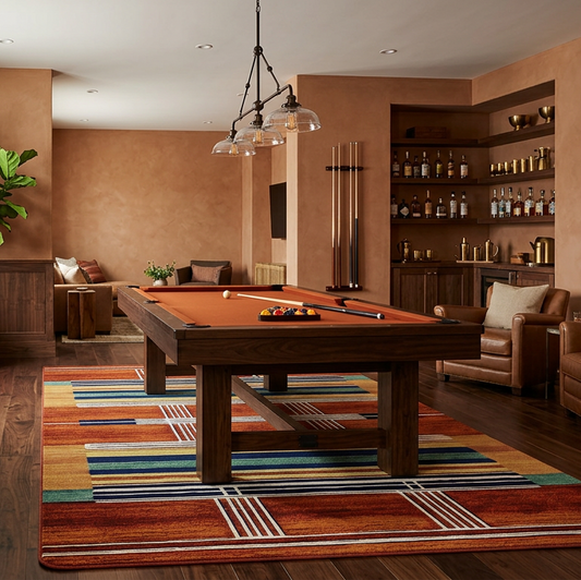 A warm, upscale interior room featuring a wooden ppool table centered on a colorful striped rug, with leather armchairs, built-in shelving, framed wall art, and wood paneling.