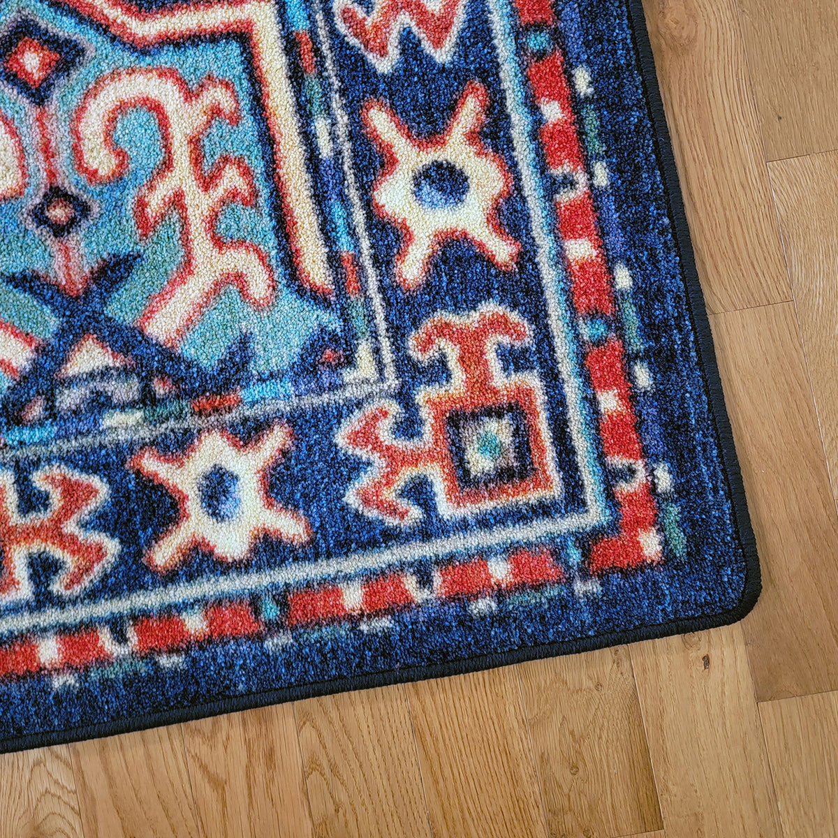 A close-up corner view of a patterned area rug on a light wood floor. The rug has a deep blue base with red, cream, and light blue geometric and ornamental motifs, finished with a dark border.