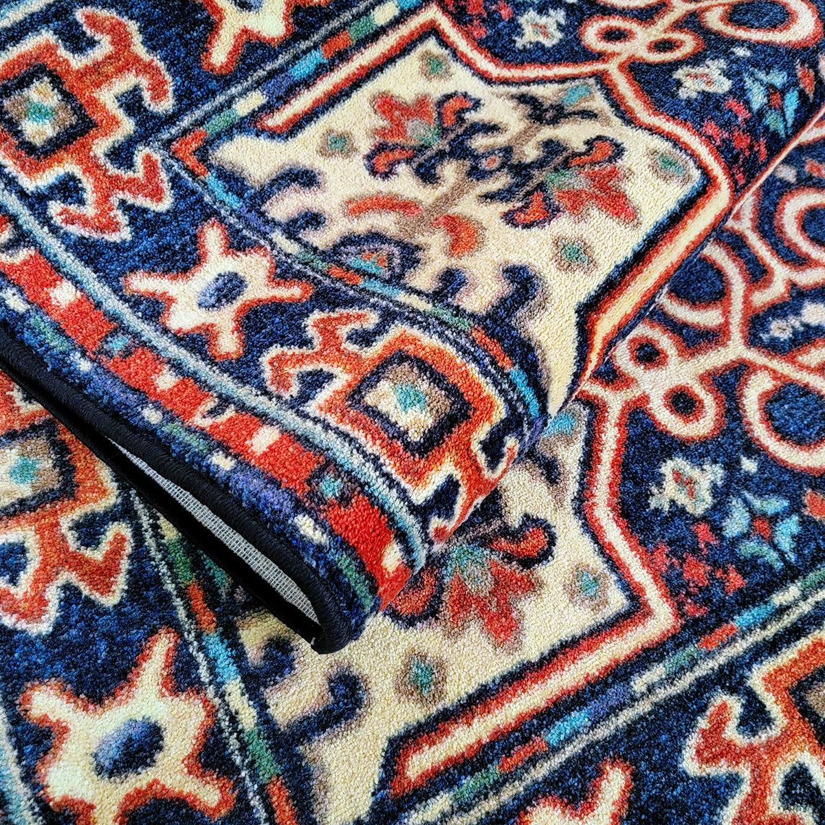 A close-up of a folded area rug with a traditional geometric pattern in navy blue, cream, red, and light blue, showing the plush pile texture and the black backing.