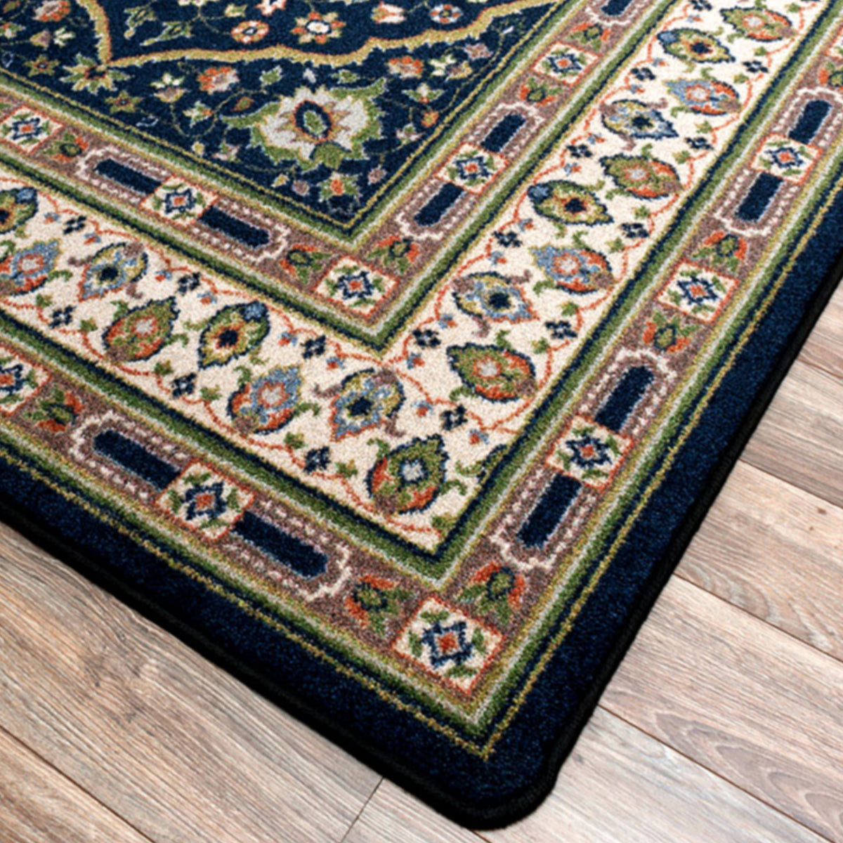 Night Garden area rug: a close-up corner view of a patterned area rug laid on a wood floor. The rug has a dark navy blue border, a cream center field, and ornate floral motifs in green, orange, and blue.