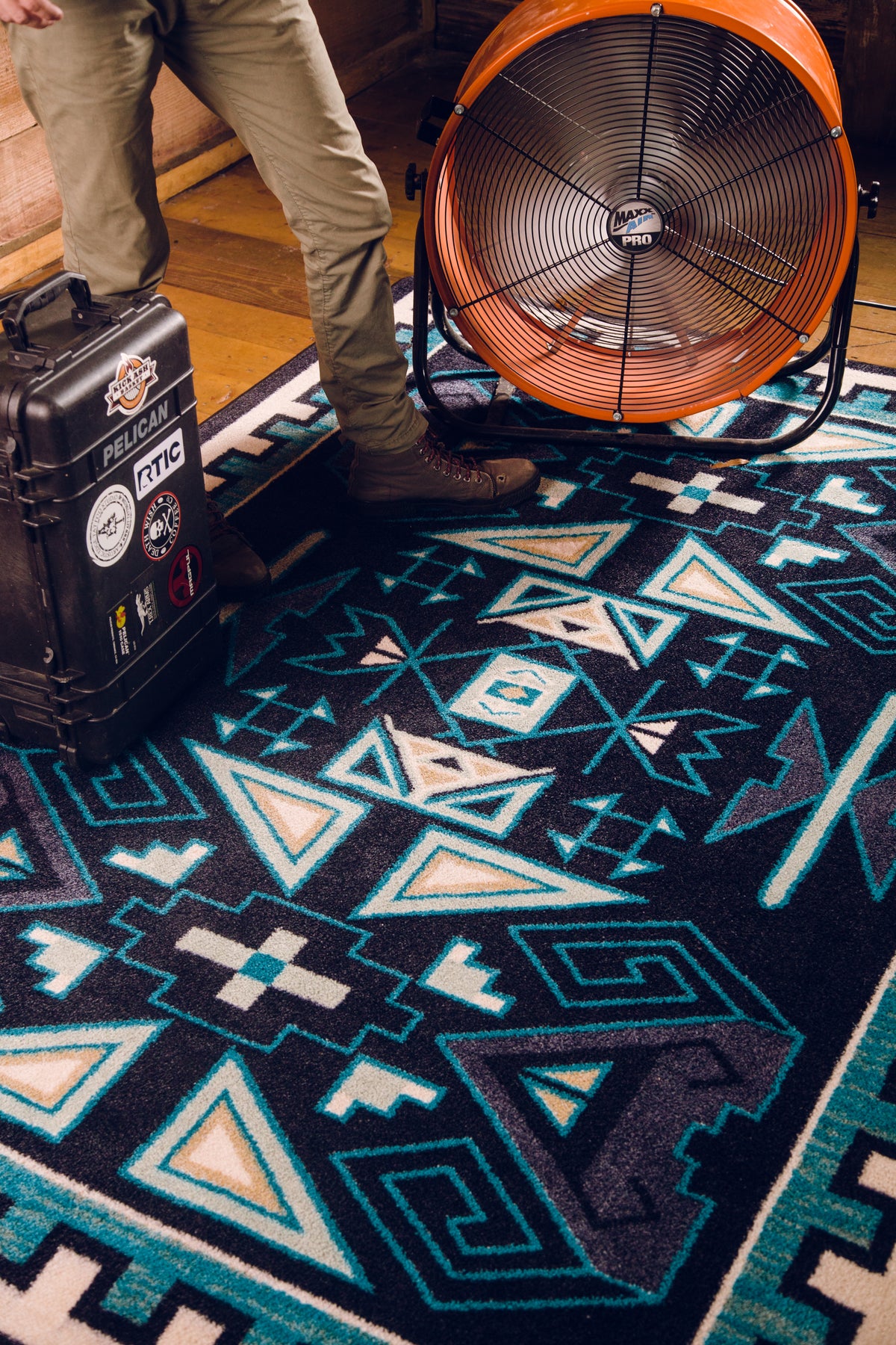 A close-up of a Southwestern-style area rug with a geometric pattern in black, teal, cream, and gray. A person's legs and boots are visible standing on the rug next to a large orange floor fan and a black…