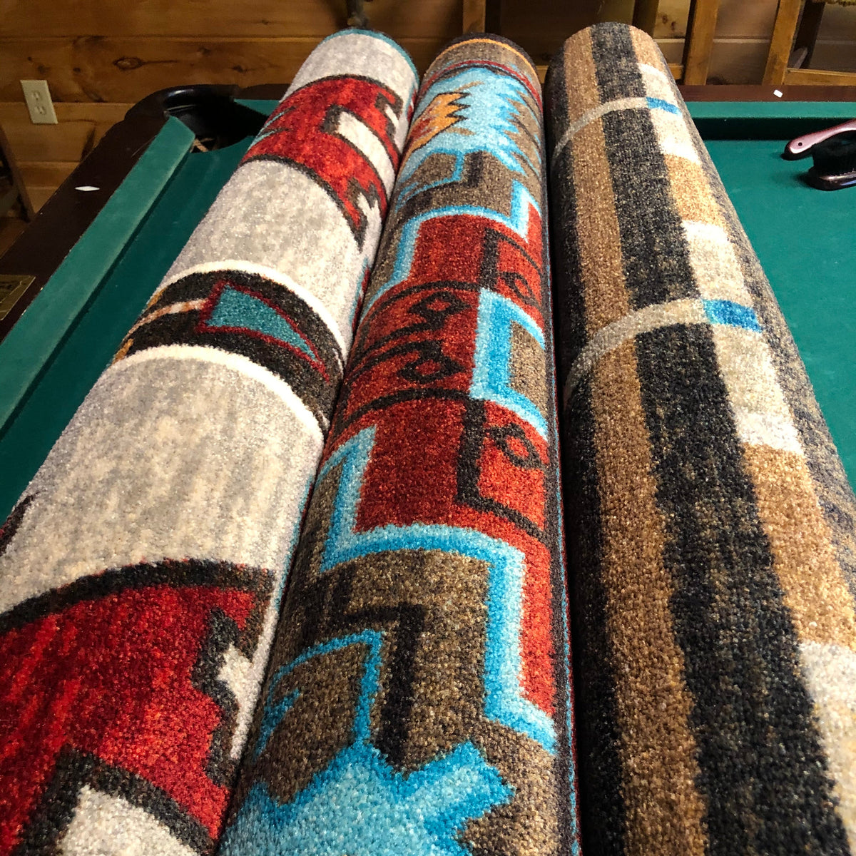 Three rolled area rugs with Southwestern geometric patterns are displayed side-by-side on a green ppool table. The rugs feature vibrant red, blue, brown, black, and beige designs.